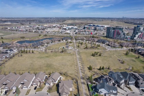 Expansive community aerial showcasing residential properties, a water feature, commercial developments, green spaces with walking paths, and distant city skyline - 2036 Ainslie Link Link, Edmonton, AB - Outdoor With View