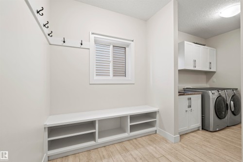 Dedicated laundry area featuring white cabinetry, a built-in bench with open storage, a window with louvered blinds, wall-mounted coat hooks, and wood-finish flooring - 2036 Ainslie Link Link, Edmonton, AB - Indoor Photo Showing Laundry Room