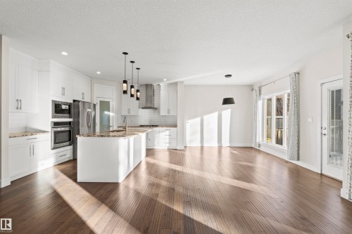 Open-concept kitchen and living area featuring wood-finish flooring, a kitchen island with pendant lighting, white cabinetry, and stainless steel appliances - 2036 Ainslie Link Link, Edmonton, AB - Indoor Photo Showing Kitchen With Upgraded Kitchen