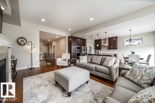 Open-concept living area featuring rich wood-finish flooring, recessed lighting, a built-in fireplace with a light-colored mantel, dark wood cabinetry in the kitchen, and stainless steel appliances - 3 8050 Orchards Green, Edmonton, AB - Indoor Photo Showing Living Room With Fireplace
