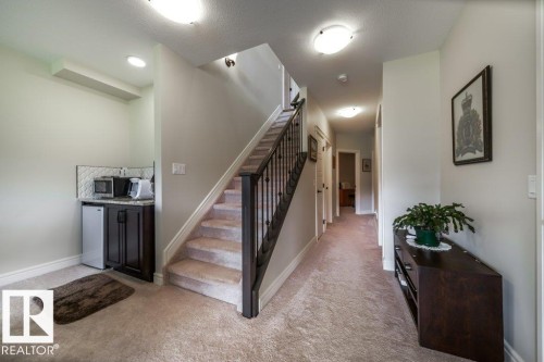 Hallway featuring a built-in wet bar with dark cabinetry, a neutral-toned carpet, a staircase with dark wood handrails and black metal spindles, and overhead flush-mount lighting - 3 8050 Orchards Green, Edmonton, AB - Indoor Photo Showing Other Room