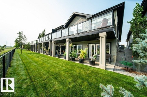 Rear exterior featuring a manicured lawn, full-length deck with glass panel railings, and an expansive lower patio - 3 8050 Orchards Green, Edmonton, AB - Outdoor With Deck Patio Veranda