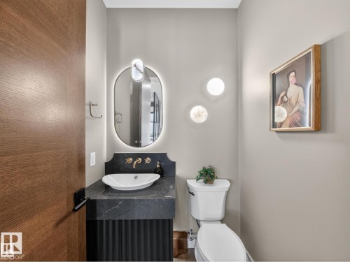Modern powder room featuring a wall-mounted oval mirror with integrated lighting, a vessel sink, a dark stone vanity top, and a fluted vanity cabinet - 7104 54 Avenue, Beaumont, AB - Indoor Photo Showing Bathroom