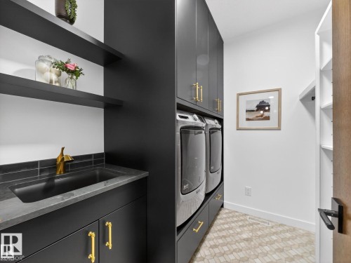 Dedicated laundry space featuring a built-in sink with a gold-tone faucet, dark cabinetry with gold-tone hardware, stacked appliances, and open shelving - 7104 54 Avenue, Beaumont, AB - Indoor Photo Showing Laundry Room