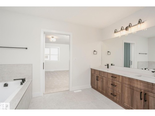 Bathroom featuring a double vanity with wood-finish cabinetry and white countertops, a large mirror with a four-light fixture, a built-in tub with tile surround, and matte black fixtures throughout - 6302 Dansereau Crescent, Beaumont, AB - Indoor Photo Showing Bathroom