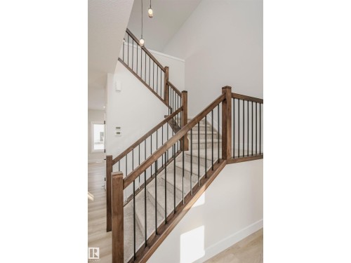 Staircase featuring wood railings with black iron balusters, light-toned carpeted treads, and pendant lighting fixtures overhead - 6302 Dansereau Crescent, Beaumont, AB - Indoor Photo Showing Other Room
