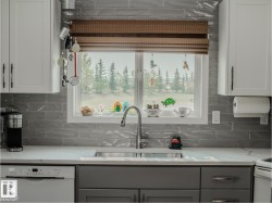Kitchen window with a woven roman shade, stainless steel double basin sink with a gooseneck faucet, light grey subway tile backsplash, white upper cabinetry, and grey lower cabinetry - 