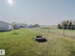 Expansive backyard featuring a built-in stone fire pit, chain link fencing, a manicured lawn, and multiple outbuildings - 