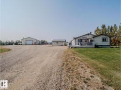 Gravel driveway with a large outbuilding featuring a roll-up door, a smaller detached outbuilding, a single-story residence with light-colored siding, and a spacious deck with white railings - 