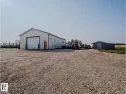 Expansive gravel lot featuring a large white outbuilding with a prominent garage door and a separate gray shed - 
