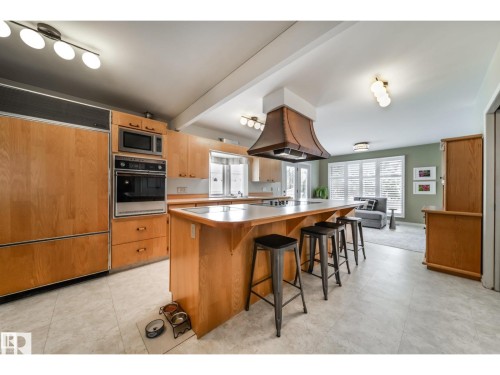 Open-concept kitchen featuring wood cabinetry, integrated oven and microwave, a center island with seating, and a decorative range hood - 10231 46 Street, Edmonton, AB 