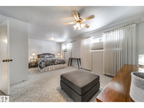 Carpeted bedroom featuring multiple windows with privacy blinds and sheer curtains, a ceiling fan with integrated lighting, and a neutral white wall finish - 10231 46 Street, Edmonton, AB 