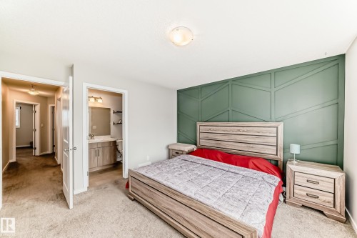 Carpeted bedroom featuring a decorative accent wall with geometric molding, a ceiling-mounted light fixture, and a doorway to a bathroom with a vanity and wall-mounted lighting - 328 Watt Boulevard, Edmonton, AB - Indoor Photo Showing Bedroom