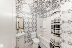 Bathroom featuring patterned wall and ceiling accents, a white vanity with integrated sink, a framed mirror, a four-bulb vanity light fixture, and a tub-shower combination with white surround - 