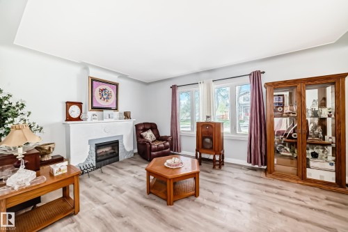 Living space featuring wood-finish flooring, a white mantel fireplace with a stone-look surround, and multiple windows with curtain rods - 11303 60 Street, Edmonton, AB - Indoor Photo Showing Living Room With Fireplace