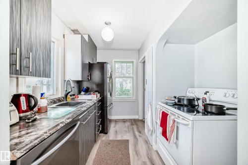 Galley kitchen featuring wood-finish flooring, dark wood-grain cabinetry with modern hardware, dark stone-look countertops, and a stainless steel dishwasher - 11303 60 Street, Edmonton, AB - Indoor Photo Showing Kitchen