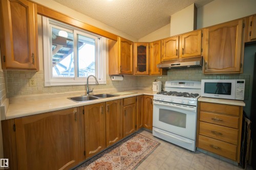 Kitchen featuring wood cabinetry, a double basin stainless steel sink, a tiled backsplash, and white appliances - 4724 38B Avenue Nw, Edmonton, AB - Indoor Photo Showing Kitchen With Double Sink