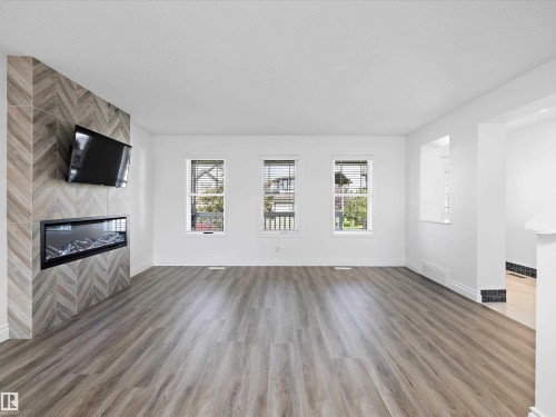 Spacious room featuring hardwood style flooring, a tiled fireplace with a modern insert, and three windows providing natural light - 9852 217 Street, Edmonton, AB - Indoor Photo Showing Living Room With Fireplace