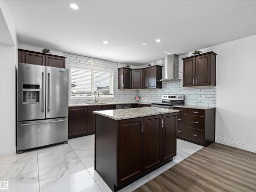 The kitchen features dark wood cabinetry, granite countertops, a stainless steel refrigerator, and a stainless steel range with a matching hood vent - 9852 217 Street, Edmonton, AB - Indoor Photo Showing Kitchen With Upgraded Kitchen