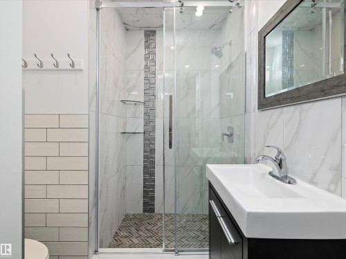 The bathroom features a sleek vanity with a rectangular sink and chrome faucet, a spacious glass-enclosed shower with a rainfall showerhead, and stylish grey and white tiled walls - 9852 217 Street, Edmonton, AB - Indoor Photo Showing Bathroom