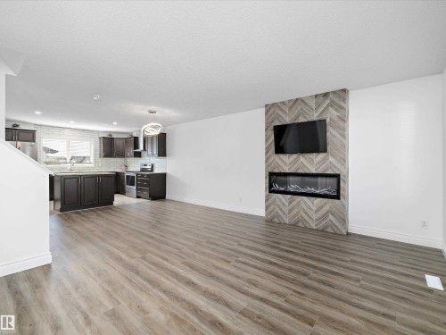 Open concept living area featuring wood-look flooring, a modern electric fireplace with a patterned tile surround, and a kitchen with dark cabinetry, stainless steel appliances, and a tiled backsplash - 9852 217 Street, Edmonton, AB - Indoor With Fireplace