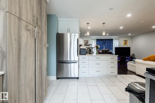 Kitchen featuring white rectangular floor tiles, a stainless steel refrigerator, and a kitchen island with white cabinetry and a light-colored countertop - 3004 68 Street, Edmonton, AB - Indoor