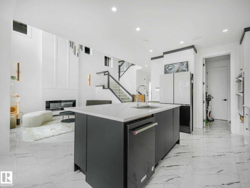 Kitchen island with a light-toned countertop and integrated sink, stainless steel dishwasher, tile flooring, built-in refrigerator, and a contemporary fireplace feature - 16307 19 Avenue, Edmonton, AB - Indoor Photo Showing Kitchen