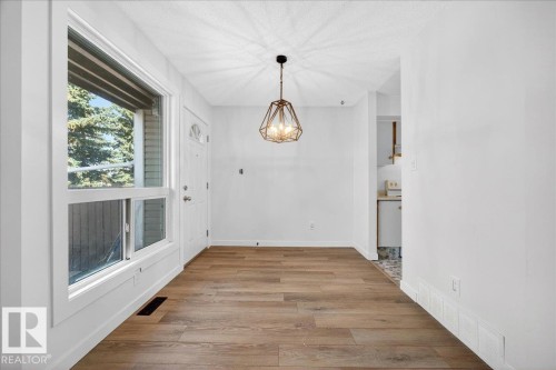 Open concept living space featuring wood-finish flooring, a modern geometric pendant light fixture, a paneled entry door with an arched window insert, and a large window with white trim - 32 1820 56 Street, Edmonton, AB - Indoor Photo Showing Other Room