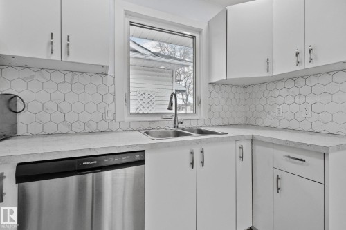 Kitchen featuring a stainless steel double basin sink, white cabinetry with silver hardware, hexagon tile backsplash, light-toned countertops, and a built-in stainless steel dishwasher - 12002 37 Street, Edmonton, AB 