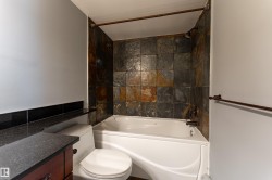 Bathroom featuring a white drop-in bathtub with a natural stone tile surround, a dark wood-finish vanity with a dark countertop, and a white toilet - 