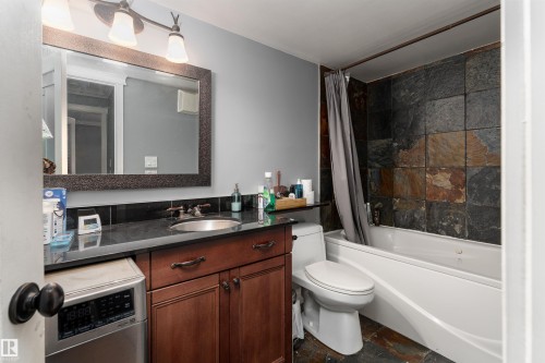The bathroom features a vanity with a dark countertop and a sink, a toilet, and a tub with natural stone tile surround - 203 10003 87 Avenue, Edmonton, AB - Indoor Photo Showing Bathroom