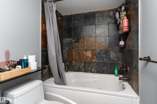 Bathroom featuring a bathtub with a shower, natural stone tile surround, and a toilet - 203 10003 87 Avenue, Edmonton, AB - Indoor Photo Showing Bathroom