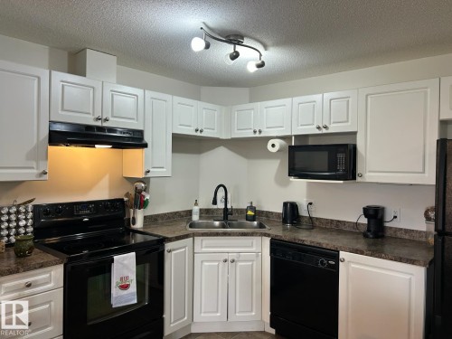 Kitchen featuring white cabinetry with brushed nickel hardware, dark countertops, a built-in oven with an electric cooktop, a mounted microwave, and a black dishwasher - 1228 9363 Simpson Drive, Edmonton, AB - Indoor Photo Showing Kitchen With Double Sink