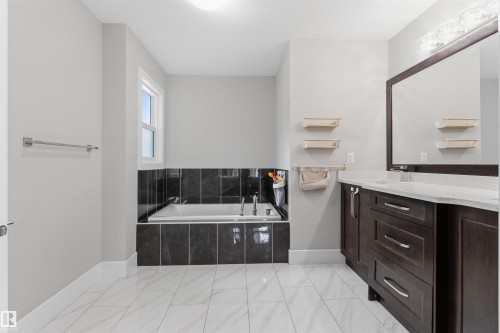 Bathroom featuring a drop-in bathtub with dark tile surround, a single window, a dark wood-finish vanity with a light countertop, a framed mirror, and light tile flooring - 3312 11 Avenue, Edmonton, AB - Indoor Photo Showing Bathroom