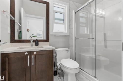 Bathroom featuring a glass-enclosed shower, a vanity with a light-toned countertop, a wood-finish cabinet with silver hardware, a framed mirror, and a window for natural light - 3312 11 Avenue, Edmonton, AB - Indoor Photo Showing Bathroom