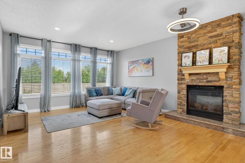 Spacious living area featuring a stacked stone fireplace with wood mantel, a series of large windows, recessed lighting, wood-finish flooring, and a contemporary ceiling fixture - 34 Valleyview Ridge, Fort Saskatchewan, AB - Indoor Photo Showing Living Room With Fireplace