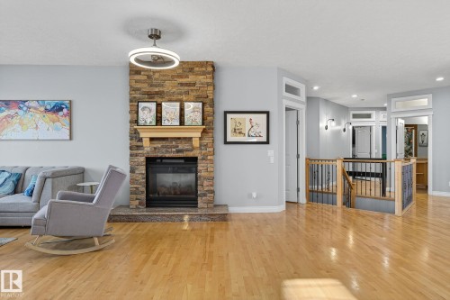 Stone-front fireplace with a wood mantle, wood-finish flooring, staircase with wood newel posts and black metal spindles, ceiling fan with integrated lighting, and wall-mounted sconces - 34 Valleyview Ridge, Fort Saskatchewan, AB - Indoor Photo Showing Living Room With Fireplace