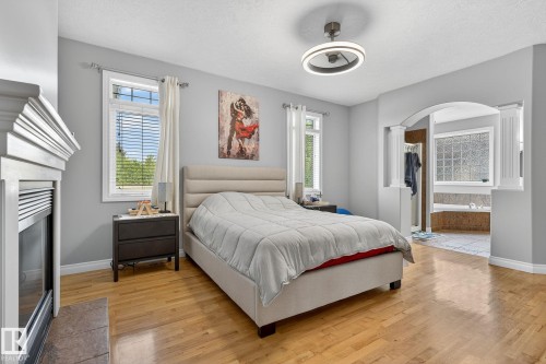 Bedroom featuring wood-finish flooring, a contemporary ceiling fan with integrated lighting, multiple windows, a decorative fireplace, and an arched entry to an adjacent space with a built-in tub - 34 Valleyview Ridge, Fort Saskatchewan, AB - Indoor Photo Showing Bedroom