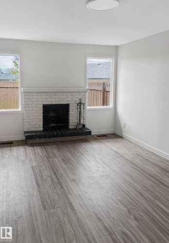 Living area featuring a white-painted brick fireplace with a dark hearth, wood-finish flooring, two large windows, and a flush-mount ceiling light - 13833 24 Street W, Edmonton, AB - Indoor Photo Showing Living Room With Fireplace