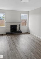 Living area featuring a white-painted brick fireplace with a dark hearth, wood-finish flooring, two large windows, and a flush-mount ceiling light - 