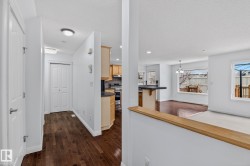 Entryway with dark wood-finish flooring, white walls, and a built-in closet, opening to a kitchen with light wood cabinetry and a peninsula countertop, and a living area featuring large windows - 
