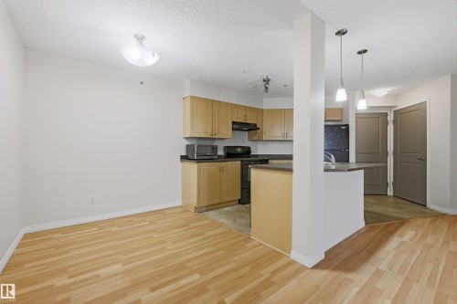 Open-concept kitchen and dining area featuring wood-finish flooring, light-toned cabinetry, dark countertops, and stainless steel appliances - 407 11453 Ellerslie Road Sw, Edmonton, AB - Indoor Photo Showing Kitchen