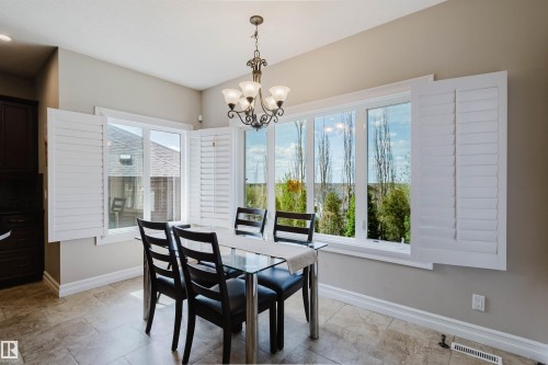 Spacious dining area featuring large windows with white louvered shutters, tile flooring, crown molding, and a decorative chandelier - 3412 Macneil Link, Edmonton, AB 