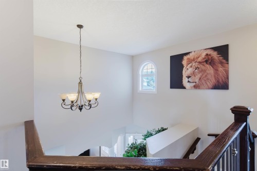 Stairwell featuring a dark wood newel post and handrail, a decorative chandelier with frosted glass shades, and an arched window with mullions - 3412 Macneil Link, Edmonton, AB 