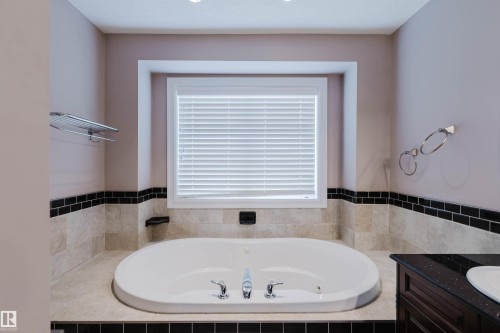Built-in soaking tub with chrome fixtures, set within a tiled surround featuring light-toned stone and a black subway tile accent - 3412 Macneil Link, Edmonton, AB 