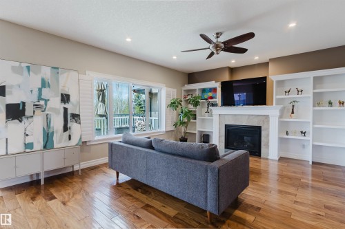Living area featuring wood-finish flooring, a fireplace with a stone surround and white mantel, built-in shelving, and a ceiling fan - 3412 Macneil Link, Edmonton, AB 