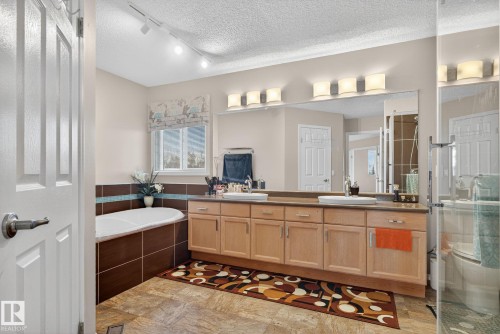 Bathroom featuring a built-in soaking tub with brown tile surround, an extended double vanity with light wood cabinetry, dual undermount sinks, and a large wall-mounted mirror - 3912 31 Street, Edmonton, AB 