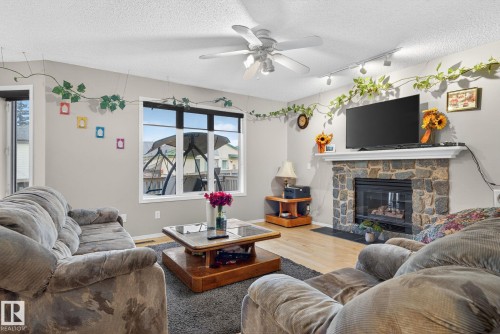 Living room featuring a stone-clad fireplace with a white mantel, light wood-finish flooring, a ceiling fan, and track lighting - 3912 31 Street, Edmonton, AB 