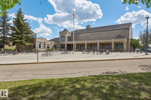 Brick building exterior with a flag pole, multiple bicycle racks, and mature evergreen trees - 18648 57 Avenue, Edmonton, AB - Outdoor With Deck Patio Veranda