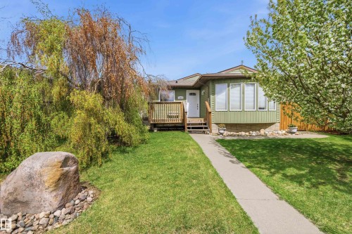 Green-sided residence featuring a front deck with lattice railing, a concrete walkway, mature trees, and a decorative large stone - 18648 57 Avenue, Edmonton, AB - Outdoor